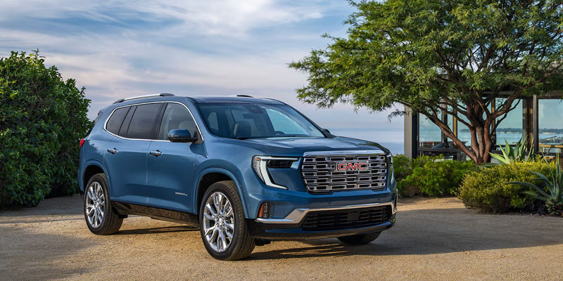 Blue GMC Acadia SUV parked outdoors near a coastal view, showcasing its front grille, headlights, and sleek exterior design.